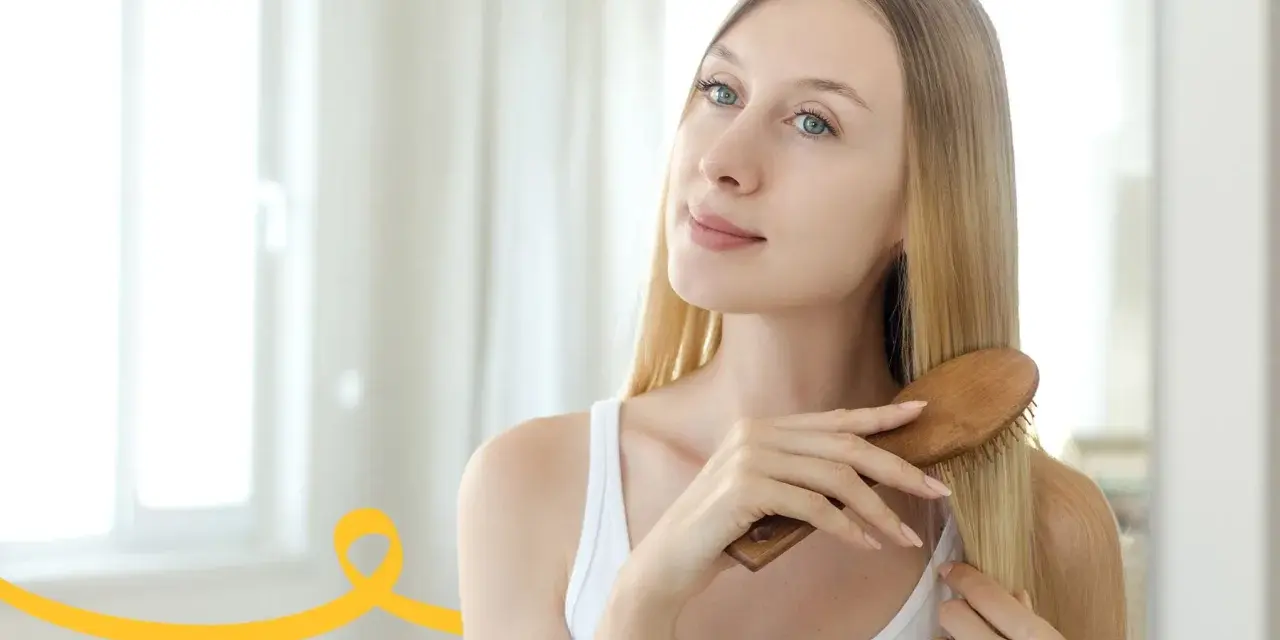 Kobieta z długimi blond włosami szczotkuje je drewnianą szczotką, dbając o oczyszczanie skóry głowy.