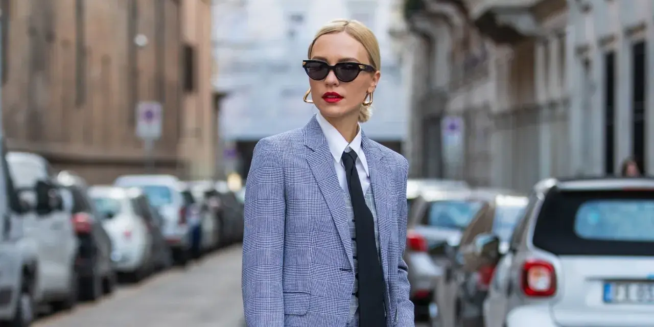 Estilo tomboy que es: mujer con traje a cuadros, camisa blanca y corbata negra, gafas de sol y labios rojos, posando en la calle.