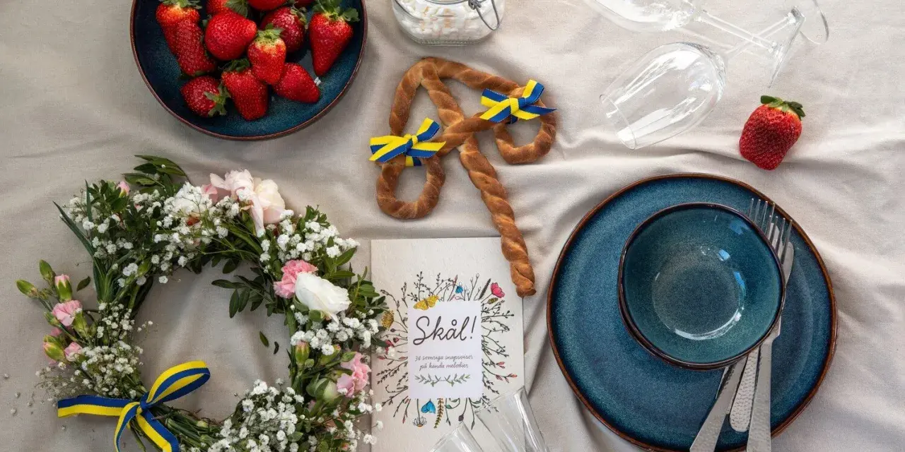 En midsommardukning med jordgubbar, blomsterkrans, en pretzel med svenska flaggor och en "Skål!"-bok. Ett personligt ljus för firandet.