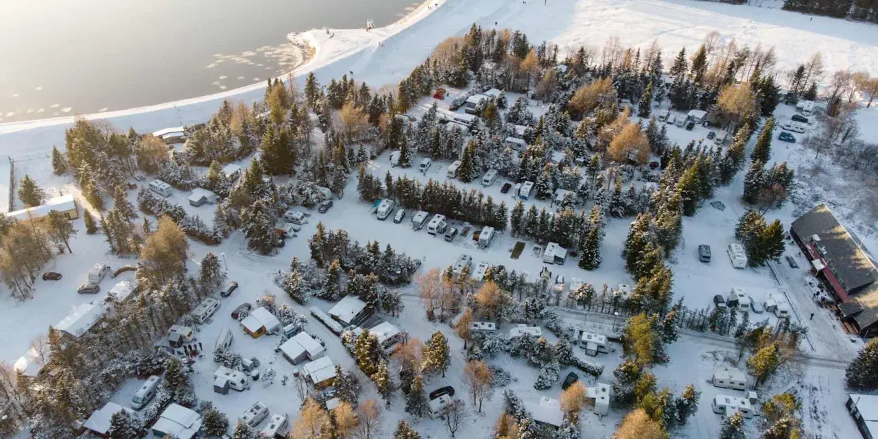 Wintercamping im Erzgebirge: Wohnmobile und Wohnwagen stehen im Schnee auf einem Campingplatz am See, umgeben von verschneiten Bäumen.