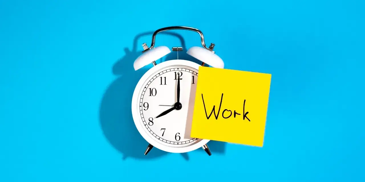 Reloj despertador blanco con nota amarilla que dice "Work", recordando la importancia de la jornada laboral.