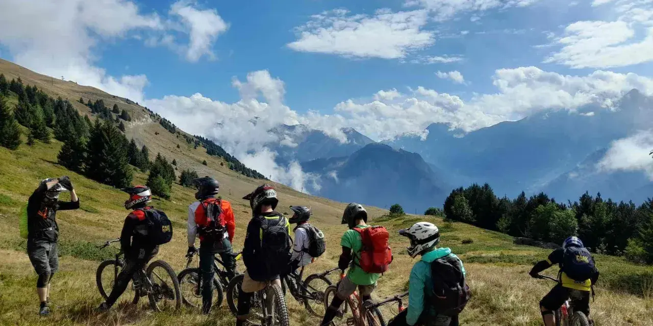 Groupe d'ados en colonie VTT descente, prêts à dévaler les pentes montagneuses sous un ciel bleu éclatant.