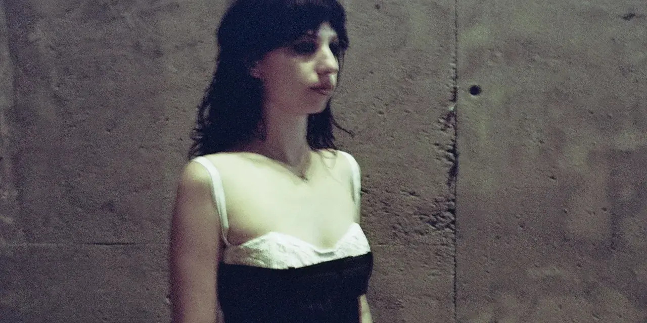 Stella Rose Gahan w czarnej sukience z białym gorsetem, stoi przy betonowej ścianie.
