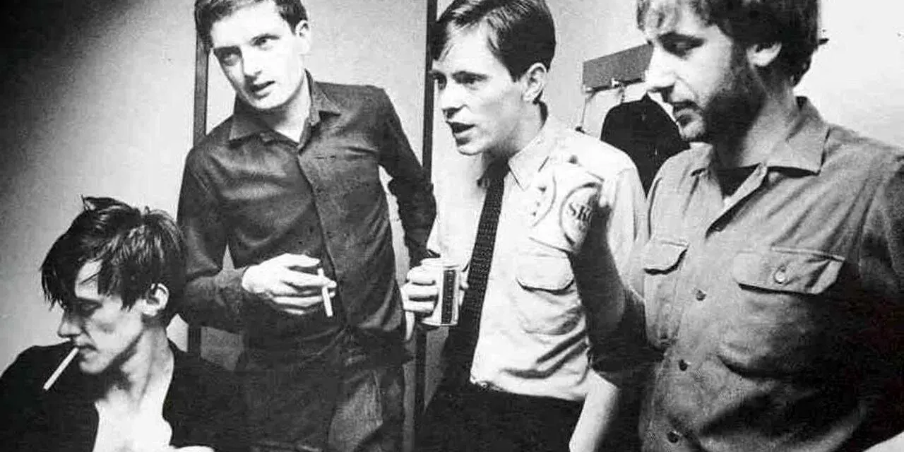 Les membres de Joy Division, Ian Curtis, Bernard Sumner, Peter Hook et Stephen Morris, posent ensemble, certains fumant, d'autres tenant des boissons.
