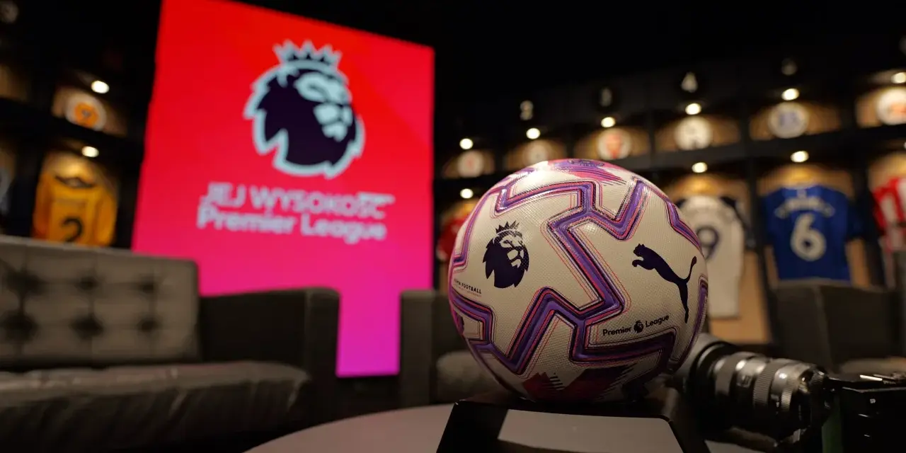 Piłka do gry w Premier League na tle ekranu z logo ligi. W tle widoczne koszulki piłkarskie.
