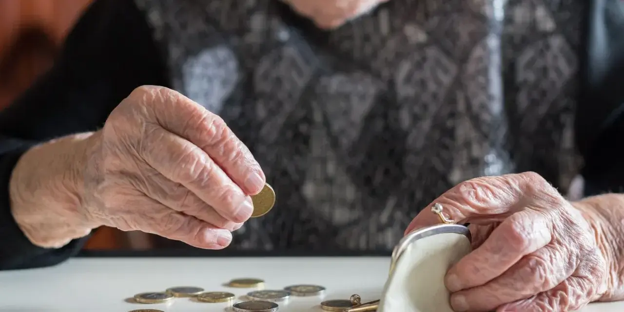 Dłoń starszej kobiety z pierścionkiem wkłada monetę do portmonetki. Rozrzucone monety na stole sugerują trudności finansowe, być może związane z emeryturą po mężu.
