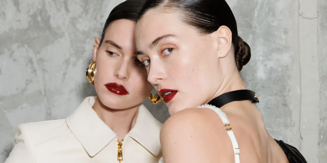 Due modelle con labbra audaci e un intenso colore rossetto, con capelli tirati indietro e orecchini dorati.