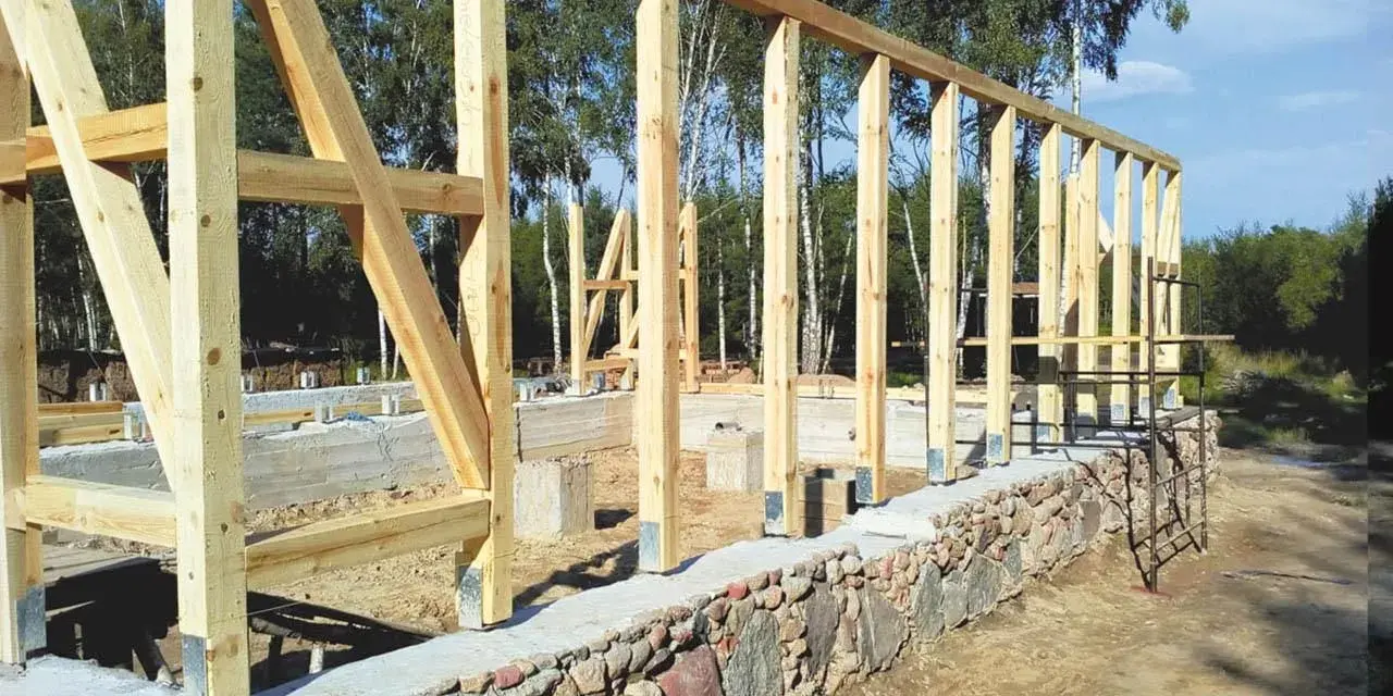 Drewniana konstrukcja domu powstaje na solidnym fundamencie z kamienia. Widać betonowe słupy i kamienny murek.