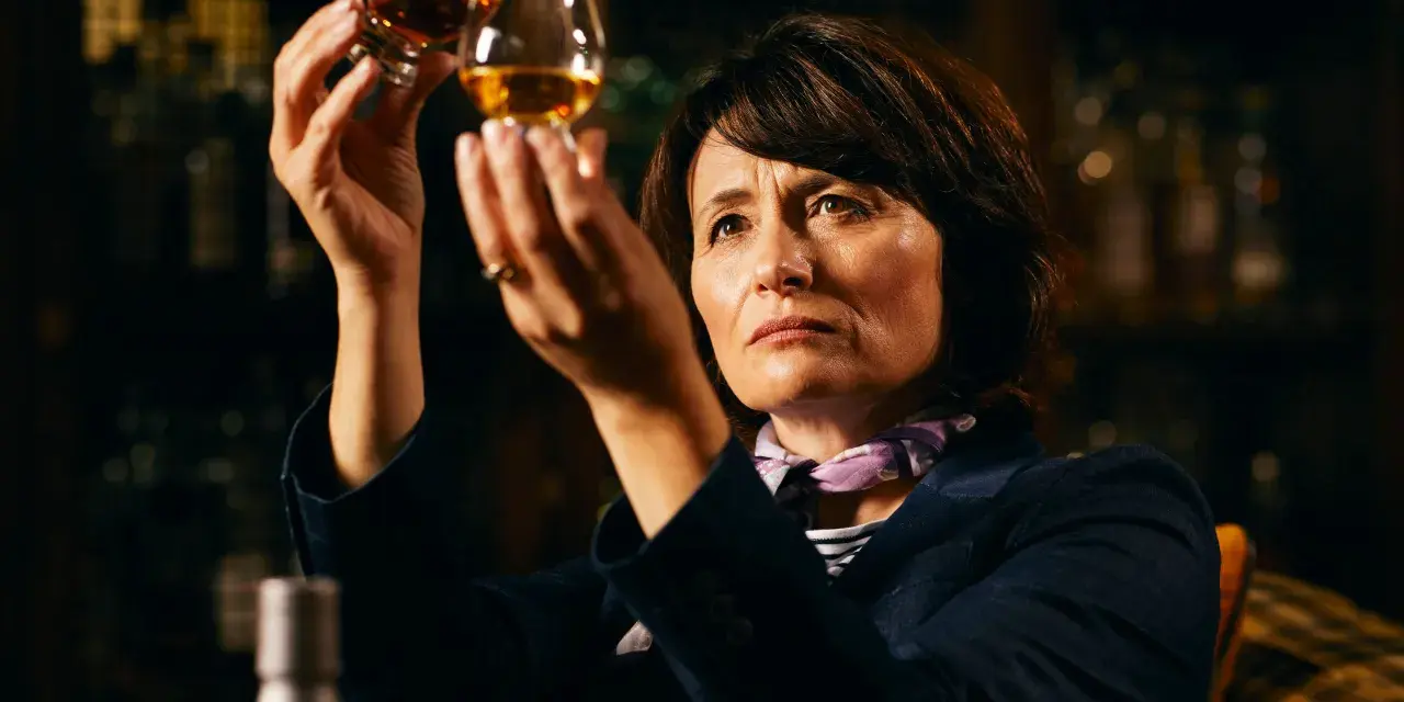 Rachel Barrie betrachtet konzentriert zwei Gläser Whisky, während sie in einer dunklen Bar sitzt.