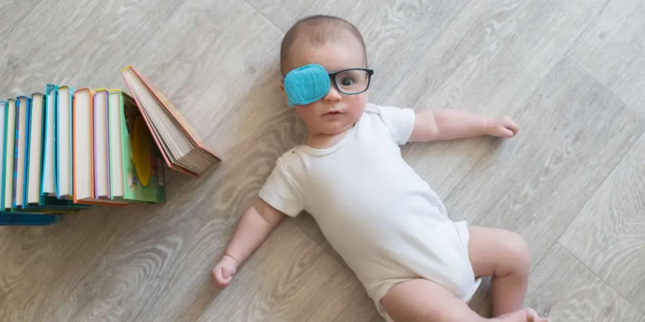 Bambino con occhiali e cerotto sull'occhio per correggere l'occhio ambliope, sdraiato vicino a una pila di libri colorati.