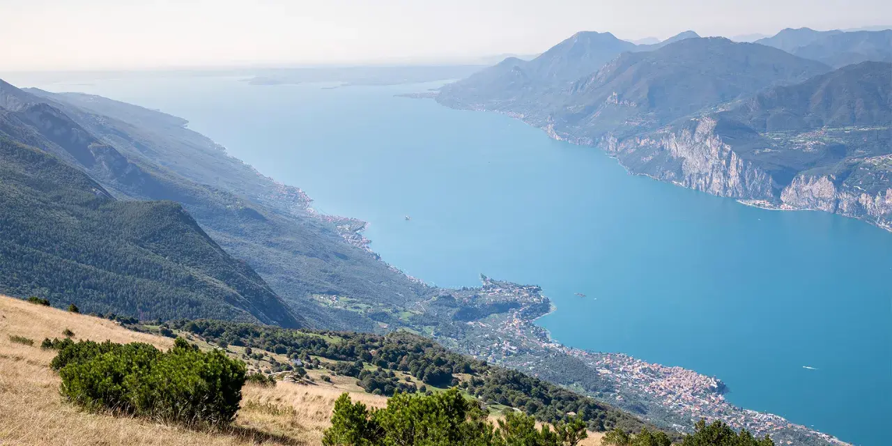 Panoramica mozzafiato del lago di Garda sponda veronese, con montagne verdi e un borgo pittoresco affacciato sull'acqua azzurra.