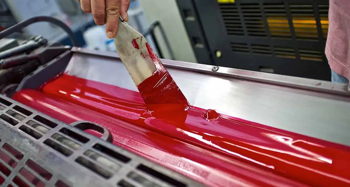 Mano esparciendo tinta roja en rodillos de imprenta, mostrando las ventajas de la imprenta para la difusión masiva de información.