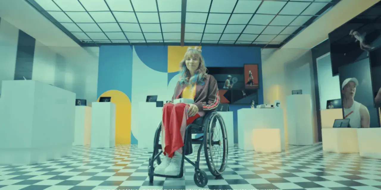 Zofia Komasa w wózku inwalidzkim, otoczona nowoczesnymi ekspozycjami, z kolorowym tłem i charakterystyczną podłogą w szachownicę.