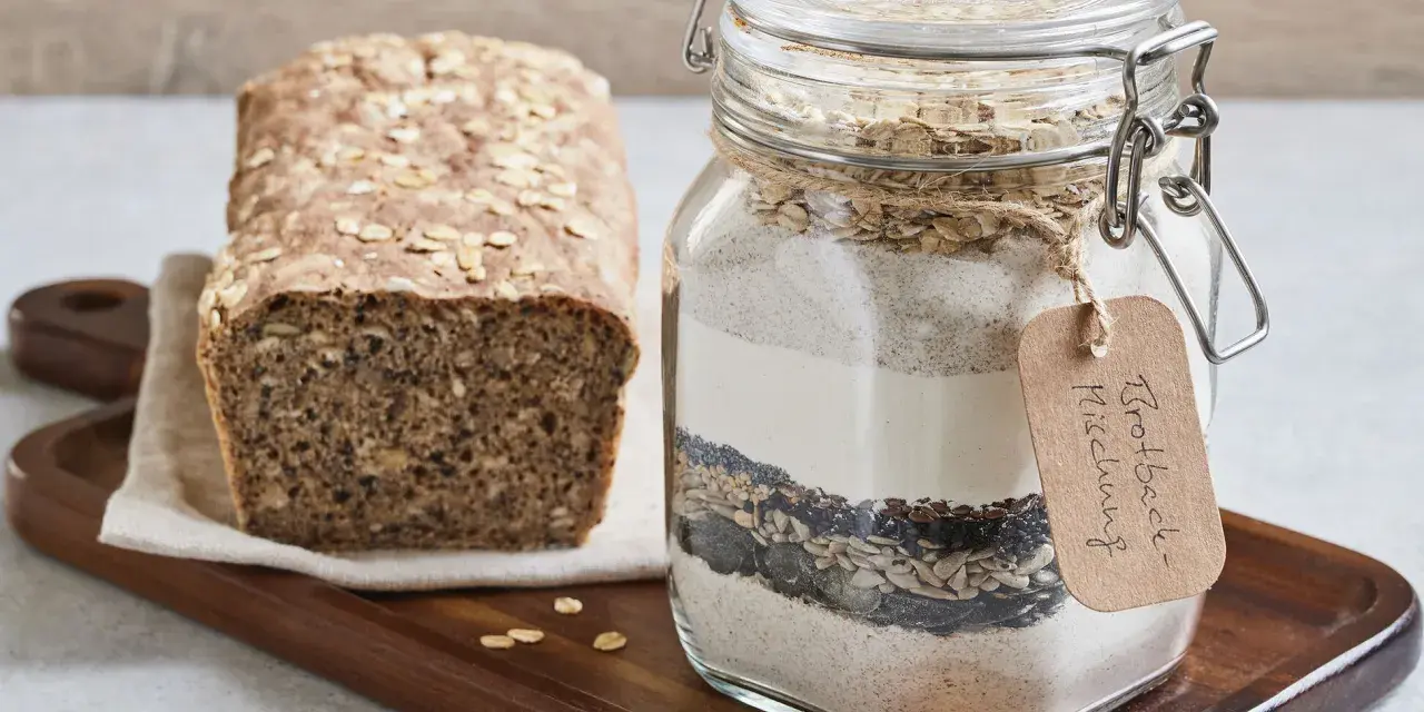 Selbstgemachte brotbackmischung im Glas, bereit zum Backen. Daneben ein frisch gebackenes Brot mit Haferflocken und Körnern.