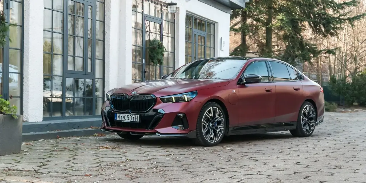 Eleganckie, matowoczerwone BMW 550e z czarnymi akcentami stoi na brukowanym podjeździe przed budynkiem z dużymi oknami.
