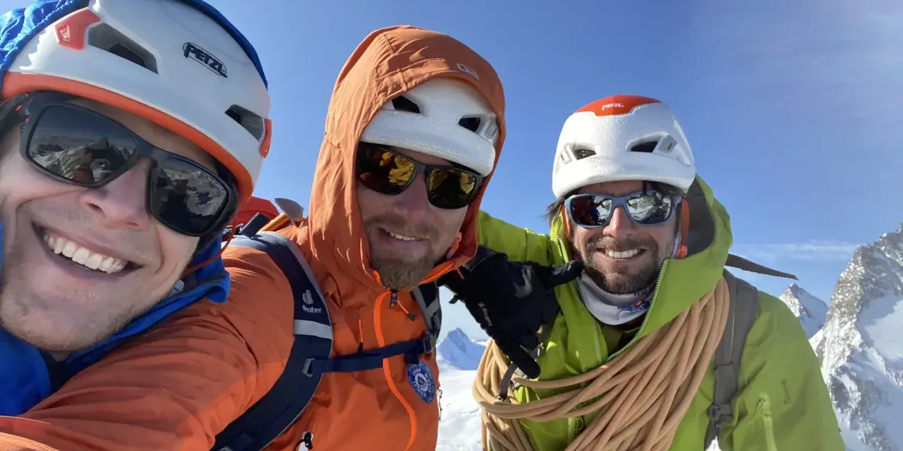 Trois alpinistes souriants au sommet, prêts à devenir guide de haute montagne à 40 ans. Corde et équipement de sécurité visibles.