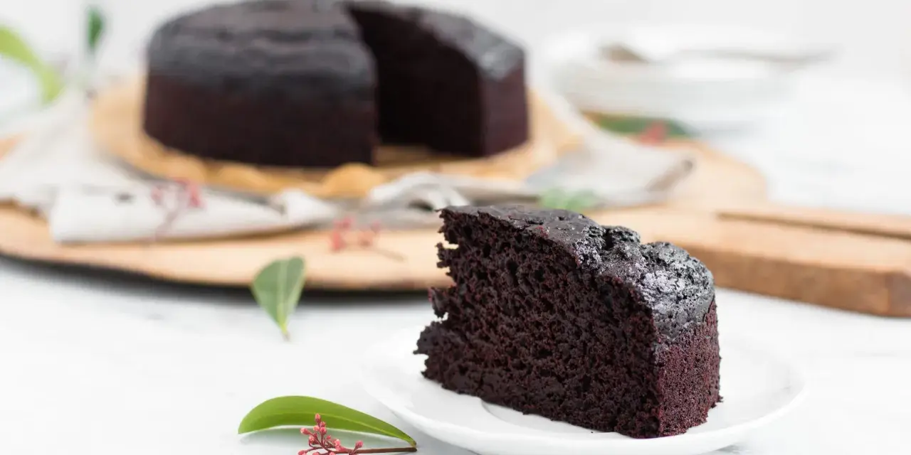 Ein saftiges Stück veganer Kuchen liegt auf einem Teller, daneben ein ganzer Schokoladenkuchen.