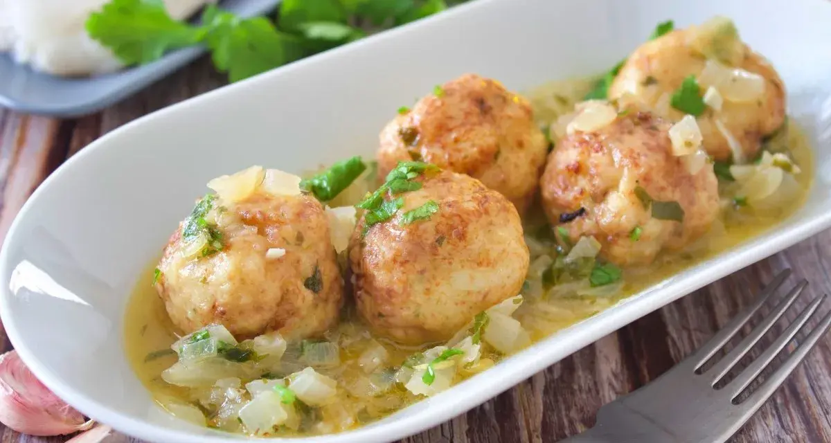 Cuatro albóndigas de merluza doradas en una salsa cremosa con cebolla y perejil, servidas en un plato blanco.