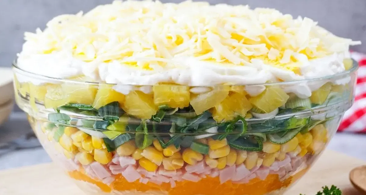 Schichtsalat-Rezept in einer Glasschüssel: Schichten aus Ananas, Lauch, Mais, Schinken, Orange, Ei und Käse.