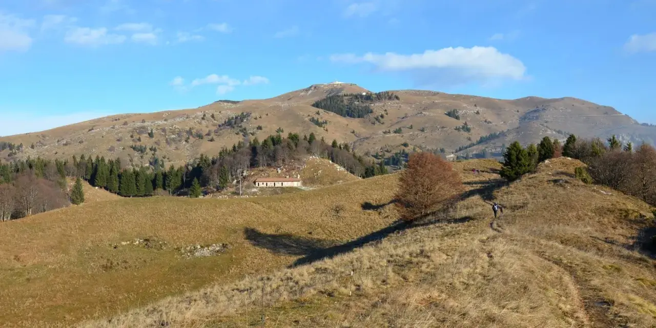 Sentieri facili sul Monte Grappa, con un escursionista che percorre un crinale erboso verso una casa isolata e la cima della montagna in lontananza.