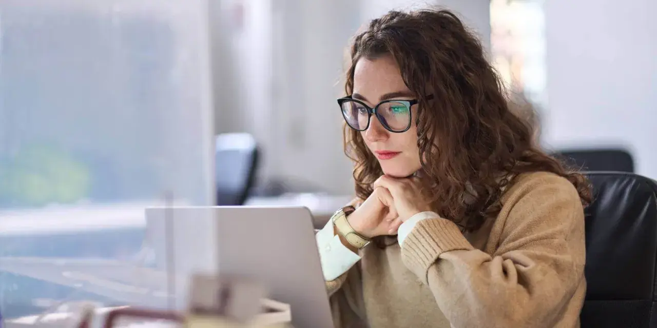 Donna con occhiali lavora al laptop, pensierosa. Potrebbe essere una dei nomadi digitali chi sono, che lavora da remoto.