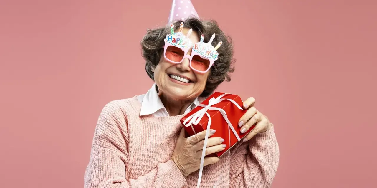 Uśmiechnięta starsza pani w okularach z napisem "Happy Birthday" i w czapce urodzinowej trzyma prezent. To dowcipne podziękowania za życzenia!