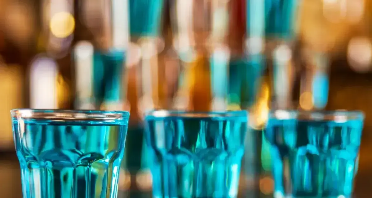 Trzy szklanki z niebieskim płynem, gotowe do wypicia jako kamikaze shot. W tle bar z butelkami.