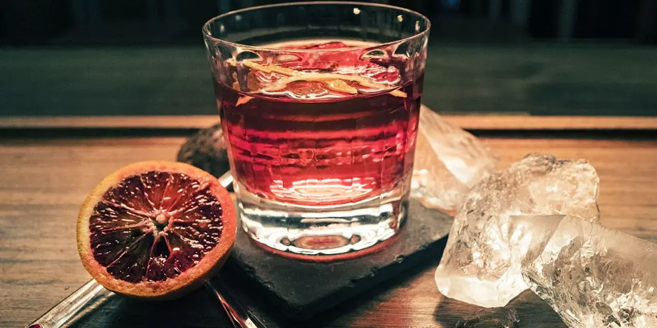 Ein Glas mit einem roten Drink, garniert mit Orangenschale, neben einer Blutorange und Eiswürfeln. Das ist Campari, ein italienischer Aperitif.