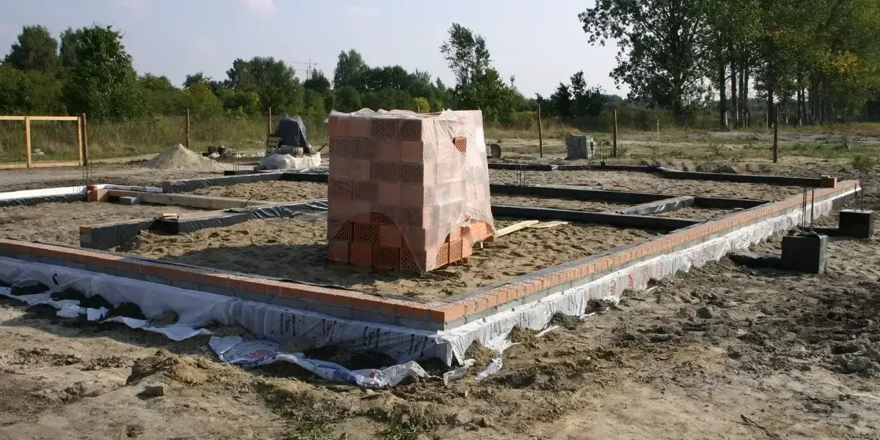 Stan zero budynku: fundamenty z cegły i betonu, piasek, folia izolacyjna, stos cegieł pod przykryciem.
