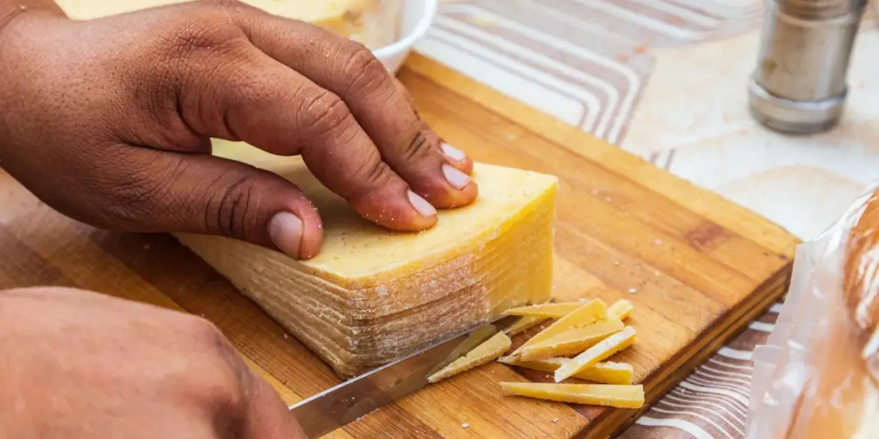 Mano cortando un bloque de queso amarillo en finas láminas. La corteza del queso se ve firme, indicando que se puede comer.