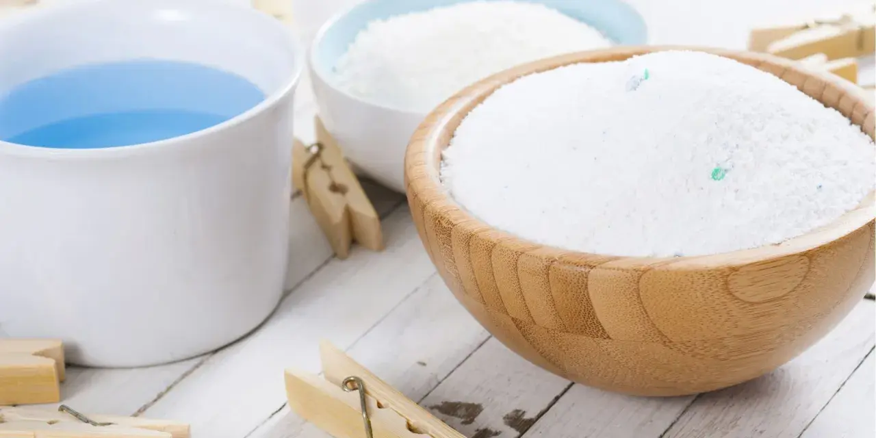 Bol en bois rempli de lessive en poudre maison, à côté d'une tasse de liquide bleu et de pinces à linge en bois.