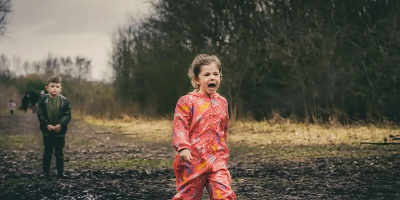 Ein 4-jähriges Kind schreit hysterisch in einem Matschfeld. Ein Junge steht im Hintergrund.