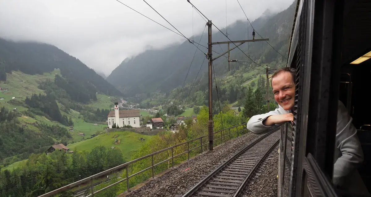 A smiling man leans out the window of the Gotthard Panorama Express, enjoying the stunning Swiss Alps scenery with a church nestled in the valley below.