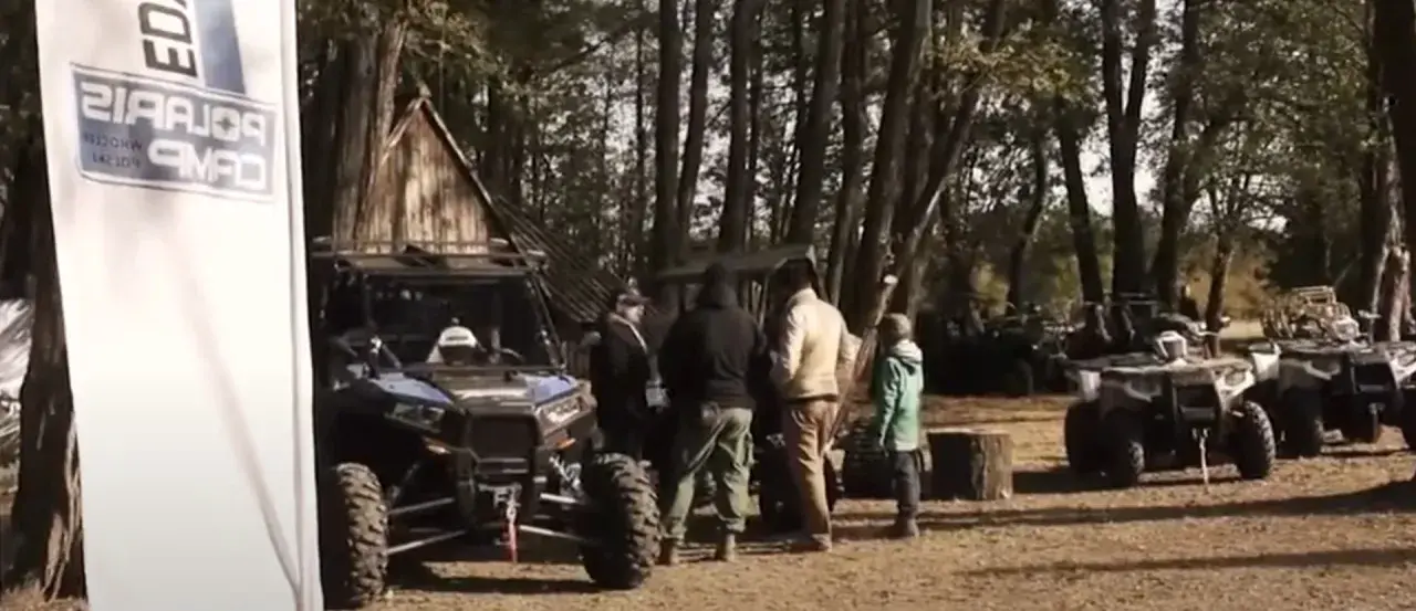 Grupa ludzi przy pojazdach terenowych podczas Polaris camping. W tle las i drewniana chata.