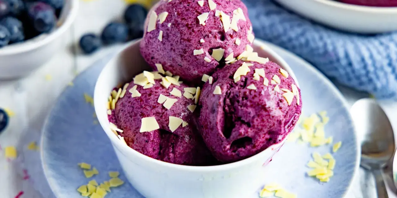 Drei Kugeln Blaubeer Eis, garniert mit weißen Schokostückchen, in einer weißen Schale auf einem blauen Teller.