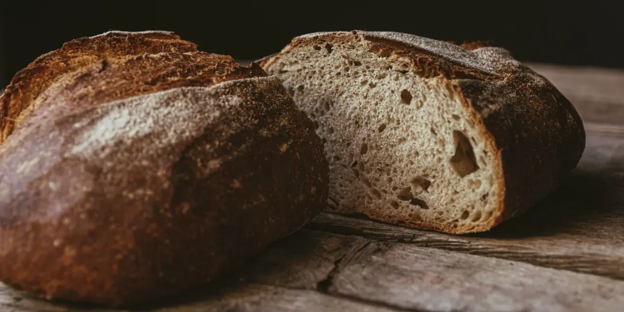 Ein rustikales, aufgeschnittenes sauerteigbrot ohne kneten liegt auf einer Holzoberfläche. Die Kruste ist dunkelbraun und knusprig, das Innere luftig und porös.