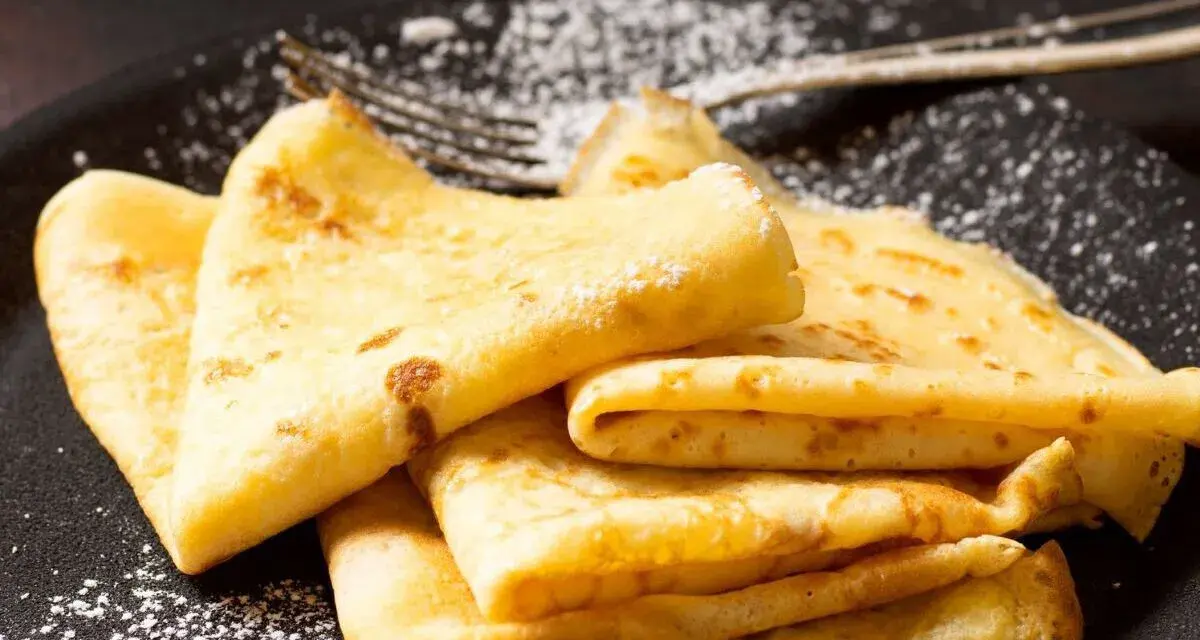 Deliciosos crepe en francés, doblados y espolvoreados con azúcar glas sobre un plato negro.