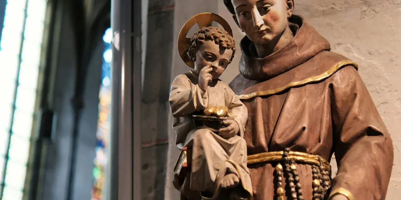 A statue shows what friars wear: brown robes, a cord belt, and a rosary. A child with a halo holds fruit.