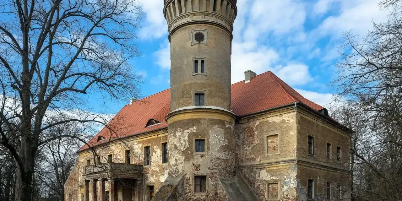 Zniszczony pałac w Osetnie z charakterystyczną wieżą i czerwoną dachówką, otoczony drzewami.