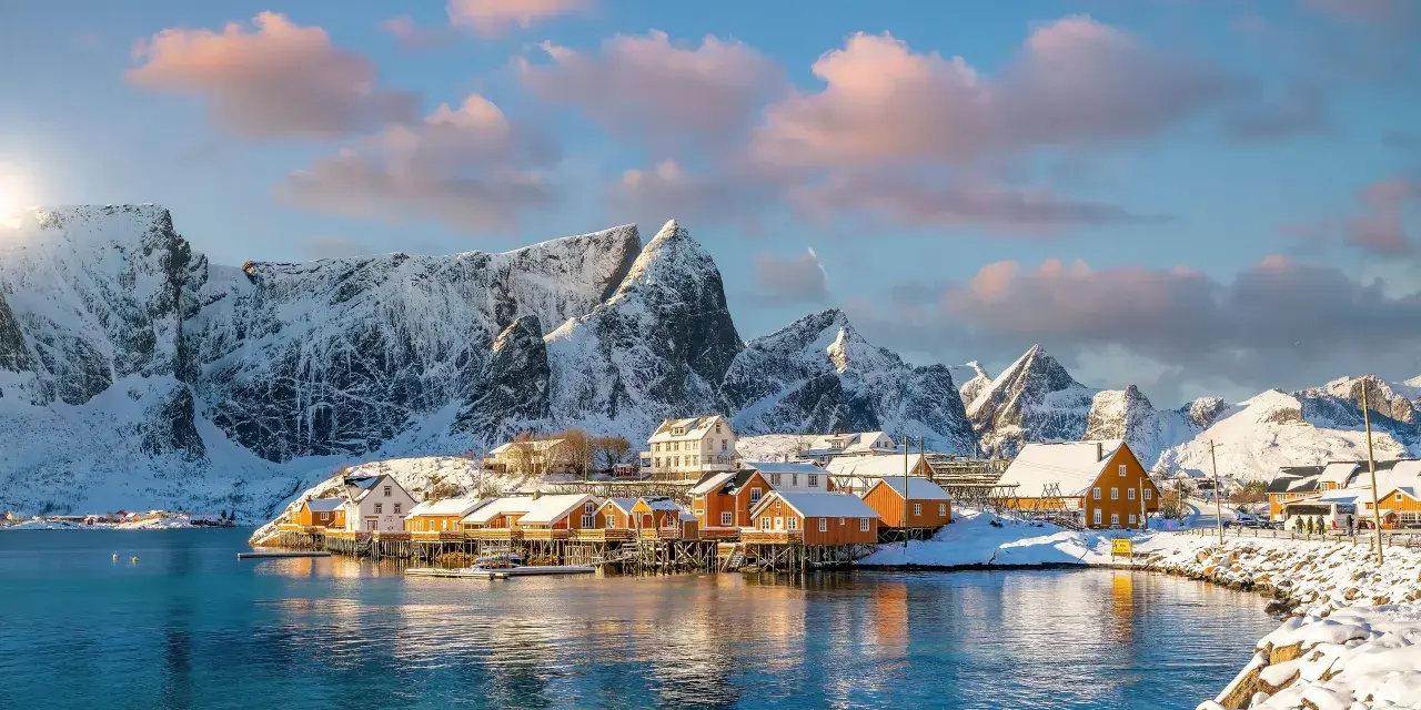 Snow-covered mountains frame a picturesque village with orange and white houses on stilts over the water, a scene from one of Europe's best winter destinations.