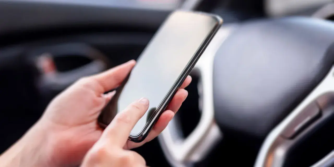 Ręce trzymają smartfon w samochodzie, obok kierownicy. Może to być pomocne, by dowiedzieć się, co to aux i jak podłączyć muzykę.