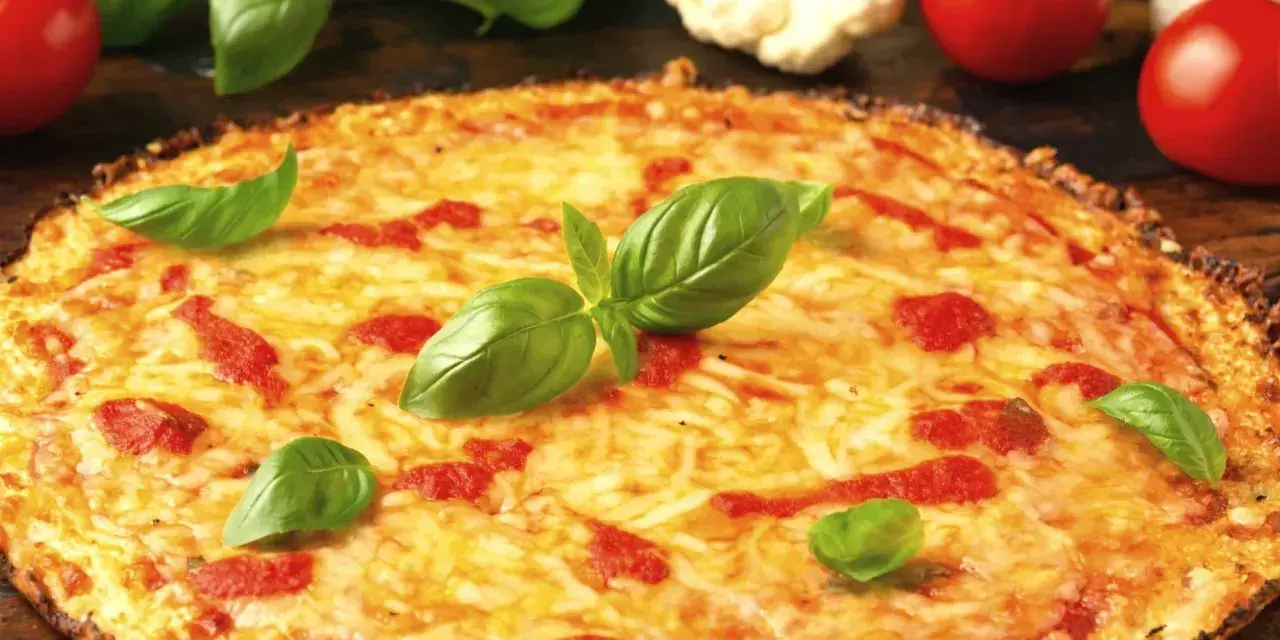 Pyszna pizza z kalafiora, z ciągnącym się serem i świeżą bazylią, otoczona pomidorami i różyczkami kalafiora.