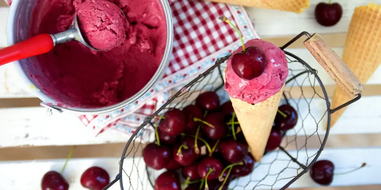 Ein Eisbecher mit Kirscheis, eine Kugel in einer Waffel, frische Kirschen und leere Waffeln.