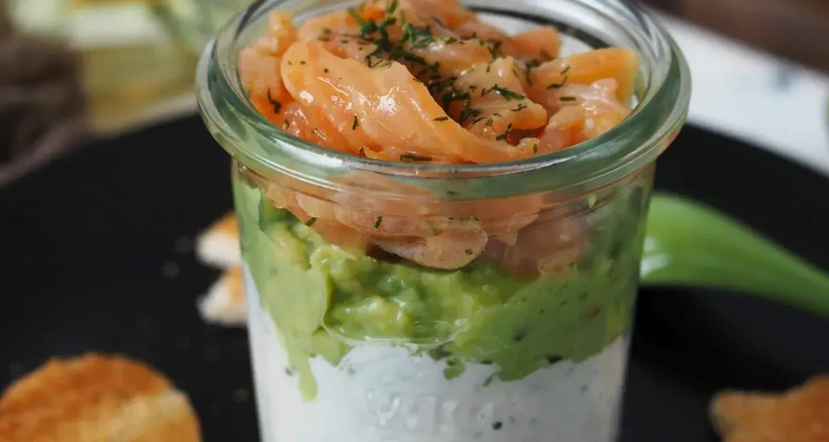 Leckerer Lachs Avocado Vorspeise im Glas, geschichtet mit cremiger Sauce und garniert mit Dill. Dazu geröstete Brotscheiben.