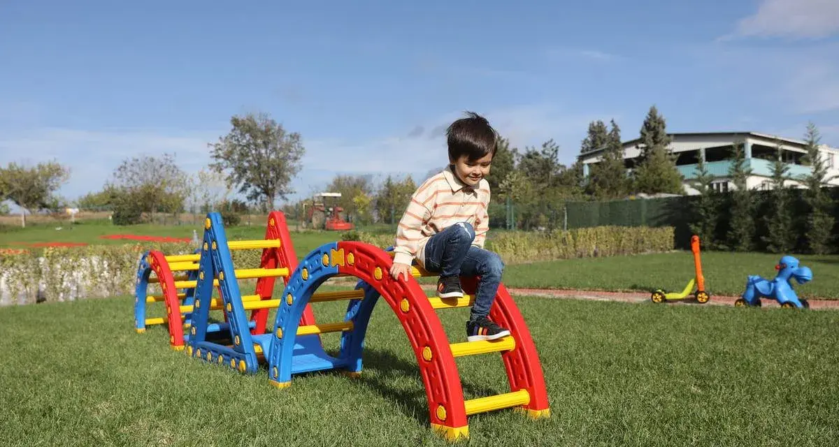 Kolorowy plac zabaw w ogrodzie to świetne atrakcje na urodziny dziecka. Chłopiec wspina się po zjeżdżalni, a obok stoi jeździk i konik na biegunach.