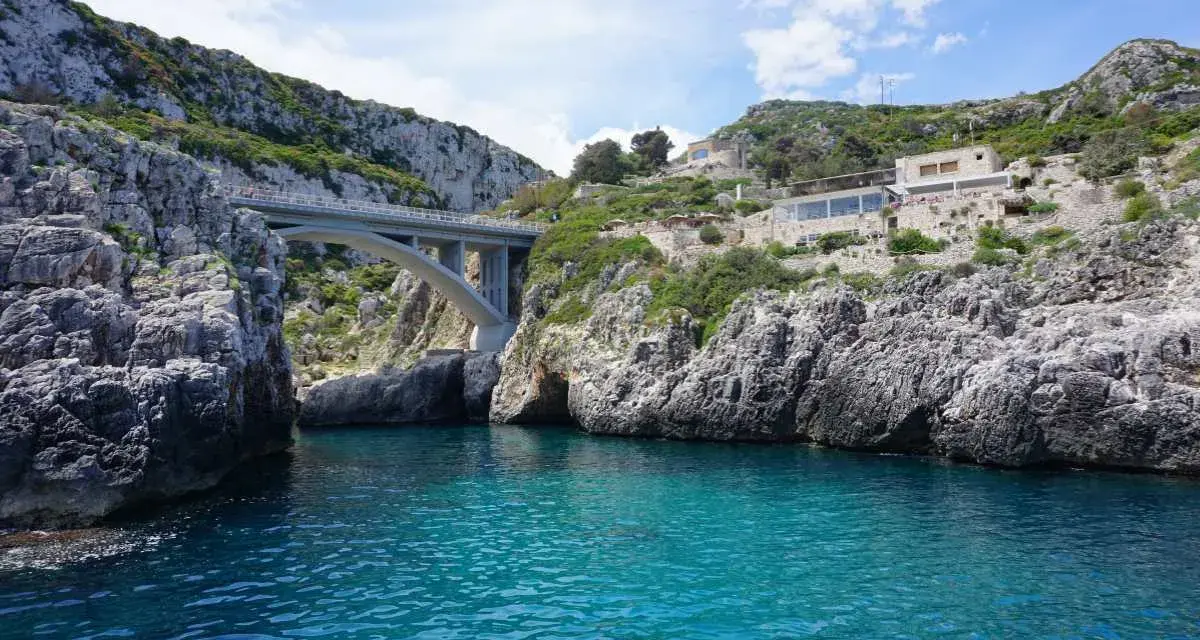 Ponte moderno su un mare cristallino, incastonato tra scogliere rocciose. Un luogo magico dove si incontrano i due mari in Puglia, con un ristorante panoramico sulla collina.