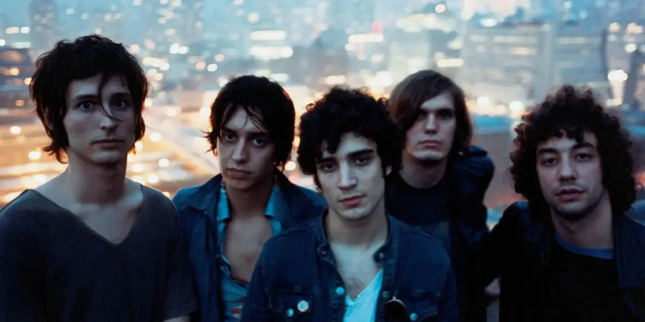 Les membres de The Strokes posent ensemble, le regard tourné vers l'objectif, avec une ville illuminée en arrière-plan.