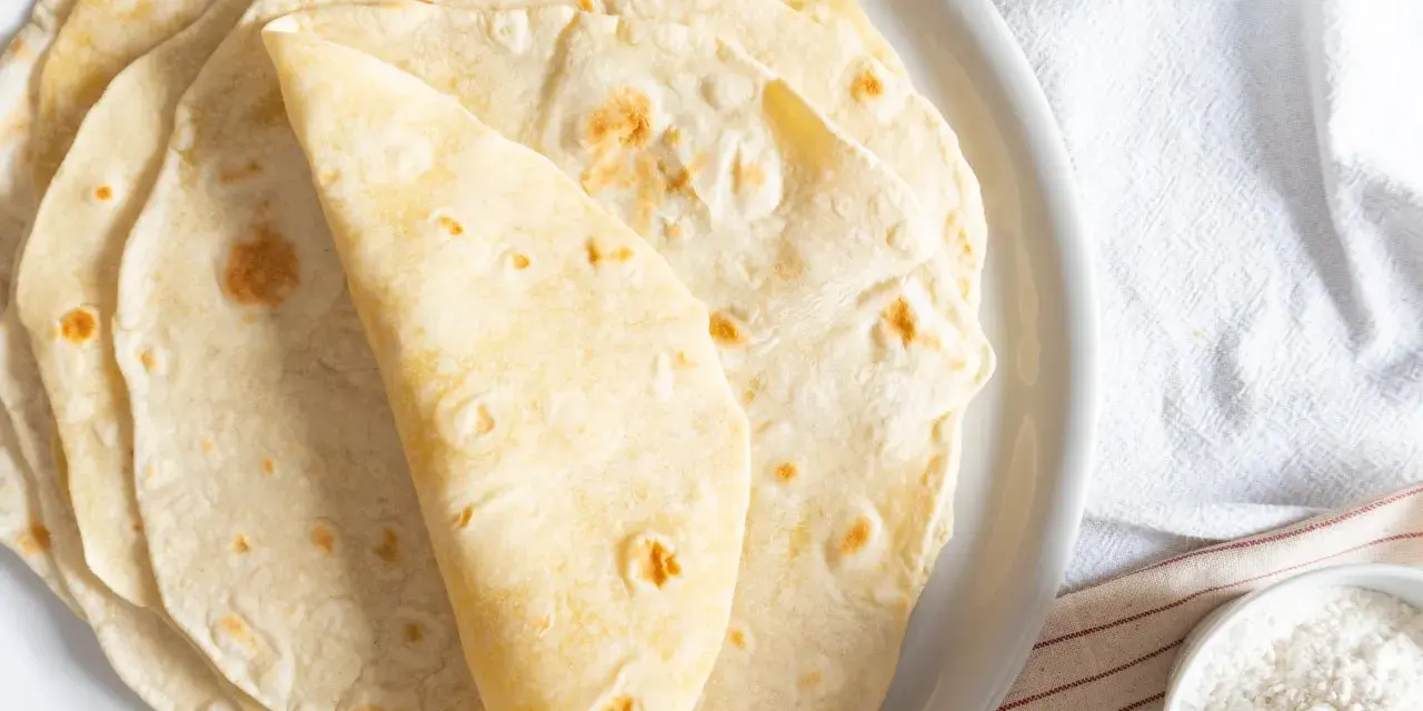 Piatto di piadine senza strutto, morbide e dorate, pronte per essere farcite. Accanto, una ciotolina d'olio e una di farina.