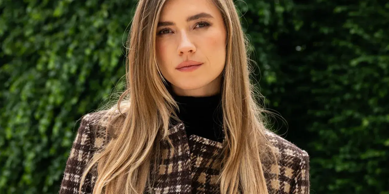 Mujer con cabello rubio y mechas invertidas, viste un abrigo de cuadros marrones y un cuello alto negro. Fondo verde frondoso.