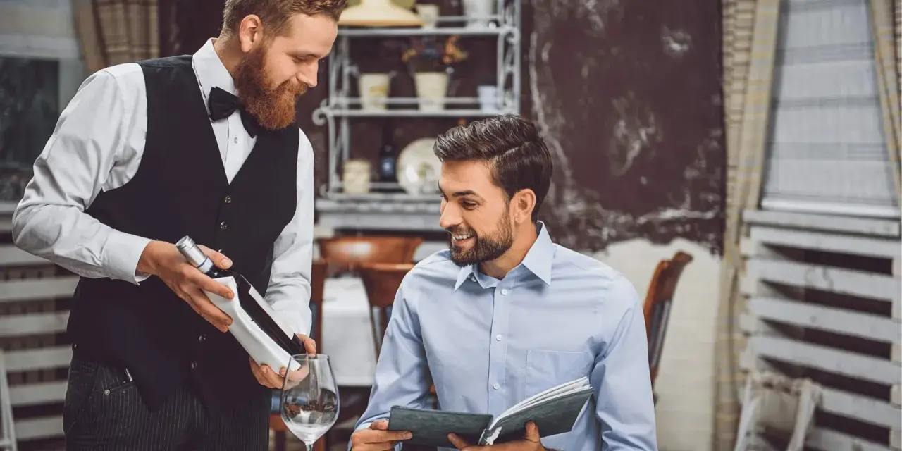 Camarero atento presenta una botella de vino a un cliente que revisa el menú. Demuestra las cualidades de un camarero: servicio, amabilidad y conocimiento.
